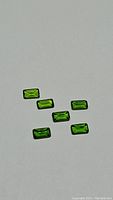 Six rectangular baguette cut vivid green chrome diopside gemstones under direct light on a white background, showing clarity and color.