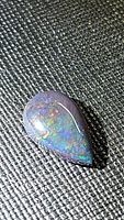 Close-up photo of pear-shaped black opal cabochon displaying play of color on textured black background.