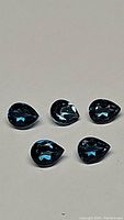 Five pear-shaped London Blue Topaz gemstones displayed on a light surface, showing deep blue color and transparency.