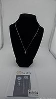 Front view of necklace displayed on black bust with certification card visible below, showing brand GRA and certification details for the 1 carat moissanite stone.