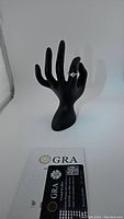 Ring displayed on black mannequin hand with certification card showing GRA label and diamond rating, confirming authenticity and stone details.