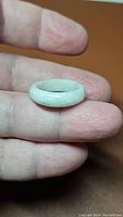 Close-up of a hand holding a smooth, rounded solid jade ring with a very light teal off white color, showing translucency and natural surface texture.
