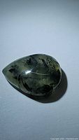Close up photo of the pear-shaped translucent green prehnite cabochon with dark rutilated inclusions, showing reflective polished surface.