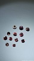 Lot of 13 natural fancy cut sapphires in shades of purple and pink arranged on a flat surface under direct light to observe color and clarity
