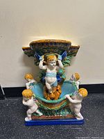 Front view of the assembled set showing the cherub wall bracket holding the plant pot with cherubs around the base, all painted in vibrant colors.