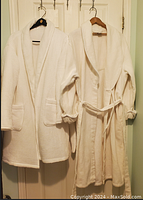 Two white bathrobes hanging on wall hooks, showing different textures and designs, one with waffle knit and elastic cuffs, other smooth with belt