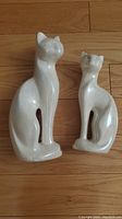 Two white porcelain cat figurines on a wooden floor, showing size difference and glossy smooth finish.