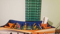 Foosball table showing playing field, yellow and blue plastic players mounted on metal rods, blue and orange side compartments, accessories in plastic bags, and booklet on the side.