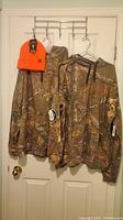 Two Realtree camouflage hoodies and an orange insulated fleece hat hanging on door hooks.