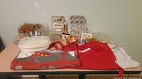 Wide view of all kitchen items displayed on table including glassware, aprons, cookbook, muffin pan, plastic containers, and utensils.