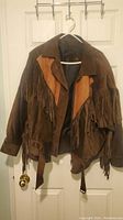 Front view of brown men's leather jacket with fringe and lighter leather accents, hanging on door.