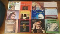 Photo showing a group of seven vinyl LP records arranged on a table, each with visible cover art. The albums include classical symphonies, operatic arias, piano and harpsichord recordings, and jazz music compilations. A book is visible among the records.
