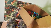 A cardboard box filled with assorted multicolored LEGO Duplo blocks beside bags containing toy figures and a colorful play mat.