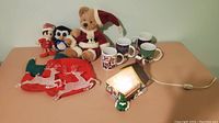 Image showing all items including the light up village house, four Christmas mugs, three plush toys (Santa bear, penguin, elf), and two red Christmas stockings with white reindeer design.