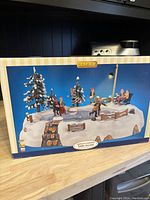 Boxed Lemax holiday-themed lighted table accent showing winter scene with snow covered trees, musicians, pond area and light post