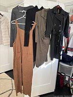 Five women's clothing items hanging on hangers including a light gray quilted vest, brown sleeveless jumpsuit, black turtleneck dress, taupe wrap dress, and black trench coat.