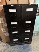 Front view of two tall, black metal filing cabinets side by side, each with four drawers with label holders and handles.