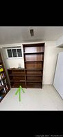Photo showing three wood bookcases side by side in a basement with a light floor and ceiling. The middle bookcase is taller and darker, the smaller one to the left has five shelves and is the subject bookcase with dimensions 41.5in high, 31.5in wide, 11in deep.
