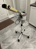 Full view of the facial steamer unit showing adjustable arm, cylindrical steaming compartment, and wheeled base on a tiled floor.