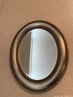 Full front view of the oval mirror showing the metallic taupe/beige frame with beaded border and clear reflective surface.