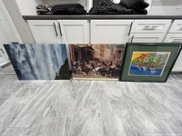 Photo of three artworks side by side: an abstract cloudscape print, a city street scene print, and a framed harbor scene painting.