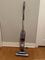 Full view of the Shark cordless stick vacuum standing upright on hardwood floor against beige wall.