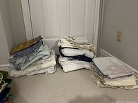 Three piles of mixed bed linens including fitted sheets, pillowcases, and top sheets in light and muted colors placed on carpet floor
