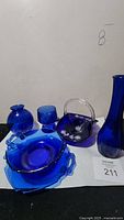 Full view of the collection showing all items: basket, plates, bowl, drinking glass, and two vases in cobalt blue glass.