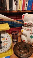 Photo showing Lucky pig cookie jar, yellow American Family Kitchen Scale, vintage Pepsi-Cola stained glass style glass, and copper pan with cowboy and bronco lid with tarnish.