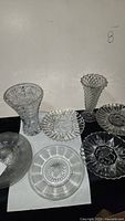 Photo showing two clear crystal vases and four crystal plates with intricate cut patterns, arranged on a white and black surface.