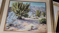 Framed oil painting depicting desert landscape with trees and mountains under blue sky.