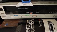 Front of Panasonic Omnivision VHS player with model and branding visible, and part of the manual and tapes below.