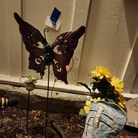 Metal purple butterfly garden stake with green flower accents, yellow artificial sunflowers, and part of blue cowboy boot visible in soil bed.