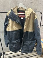 Front view of the two-tone beige and dark gray Burton snowboarding jacket hanging on a rack against wall with sherpa lining visible inside.