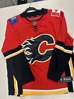 Front view of Calgary Flames red hockey jersey with black sleeves, large team logo on front, and flag patches on shoulders.