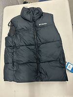Front view of new Columbia black puffer vest with logo on left chest, zipper closed, tag visible on side pocket.