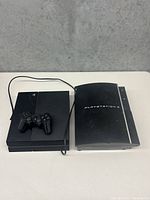 Both PlayStation 3 and 4 consoles together with a wired controller on top of the PlayStation 4 console, showing their front and top designs.