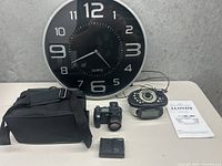 Large black quartz wall clock, vintage-style camera, black camera bag, battery charger, and Lloyd's alarm clock radio with instruction manual on table