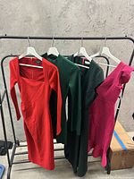 Four designer dresses hanging on a rack in front of a concrete wall. Colors include red, two shades of green, and pink.