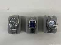 Three designer wristwatches displayed on grey cushions, front view showing faces and metal link bands