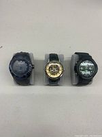 Front view of three watches displayed on grey cushions: Miykon black and blue watch, gold-tone skeleton watch, black digital-analog watch