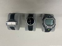 Overview of three watches on display holders: a silver clasp bracelet watch, a Calvin Klein black dial leather strap watch, and a Timex Ironman digital watch.