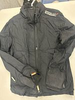 Black Superdry jacket laid flat showing front with zipper and ribbed cuffs, Japanese text logo on right chest.