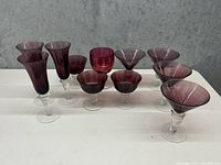 Photo of 9 cranberry glass drinking glasses in varying shapes on a light surface with grey background