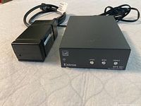 Extron MPA 601 amplifier with three front panel adjustment knobs and power adapter lying beside it on a white surface.