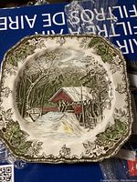 Top view of square ceramic plate featuring a red covered bridge with surrounding trees and stream, decorative green and brown floral border.