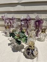 Photo showing 14 miniature coal oil lamps arranged on a white cloth. Several lamps have purple ribbon decoration and plastic wrapping. One lamp is in a small basket with green artificial leaves.