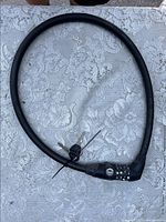 Black flexible cable bike lock arranged in a loop on a white lace fabric background, showing the combination lock mechanism.