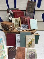 Photo showing wide assortment of antique books, mostly leather and hardbound, arranged on a wicker basket and table, including titles and black and white photos as decoration.