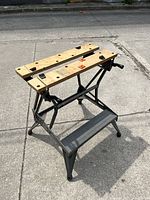 Full view of the Black & Decker Workmate folding workbench showing wooden top with multiple holes and swivel clamps, black metal frame, and front step support.